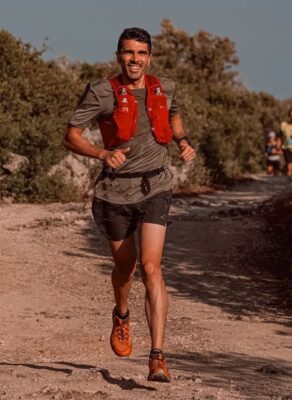 Coureur de trail courant sur un sentier ensoleillé, équipé d’un gilet rouge