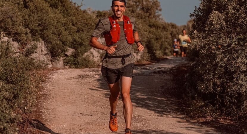 Coureur de trail courant sur un sentier ensoleillé, équipé d’un gilet rouge